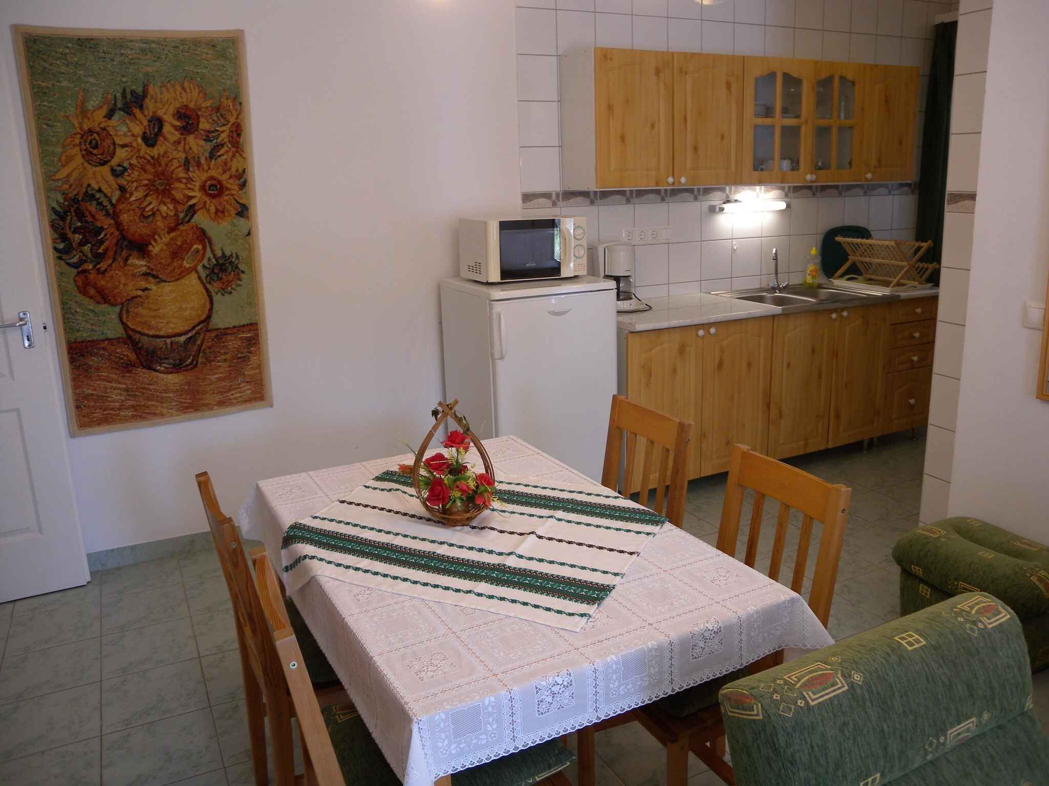 A small kitchen with wooden cabinets, a microwave, and a sink. There is a dining table with a white tablecloth and chairs. A painting of sunflowers is on the wall.