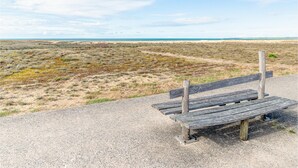 Eine hölzerne Bank an einem Kiesweg mit Blick auf eine Küstenlandschaft, spärlicher Vegetation und einem klaren Himmel.