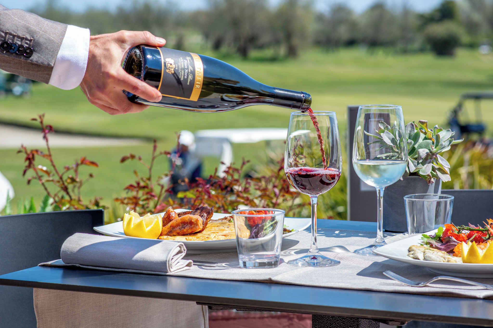 Una persona sirve vino en una copa sobre una mesa al aire libre, con platos de comida, vasos y una botella de vino.