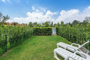 A well-maintained garden with a lawn, a hedge, and a row of plants. There are two white lounge chairs in the foreground.