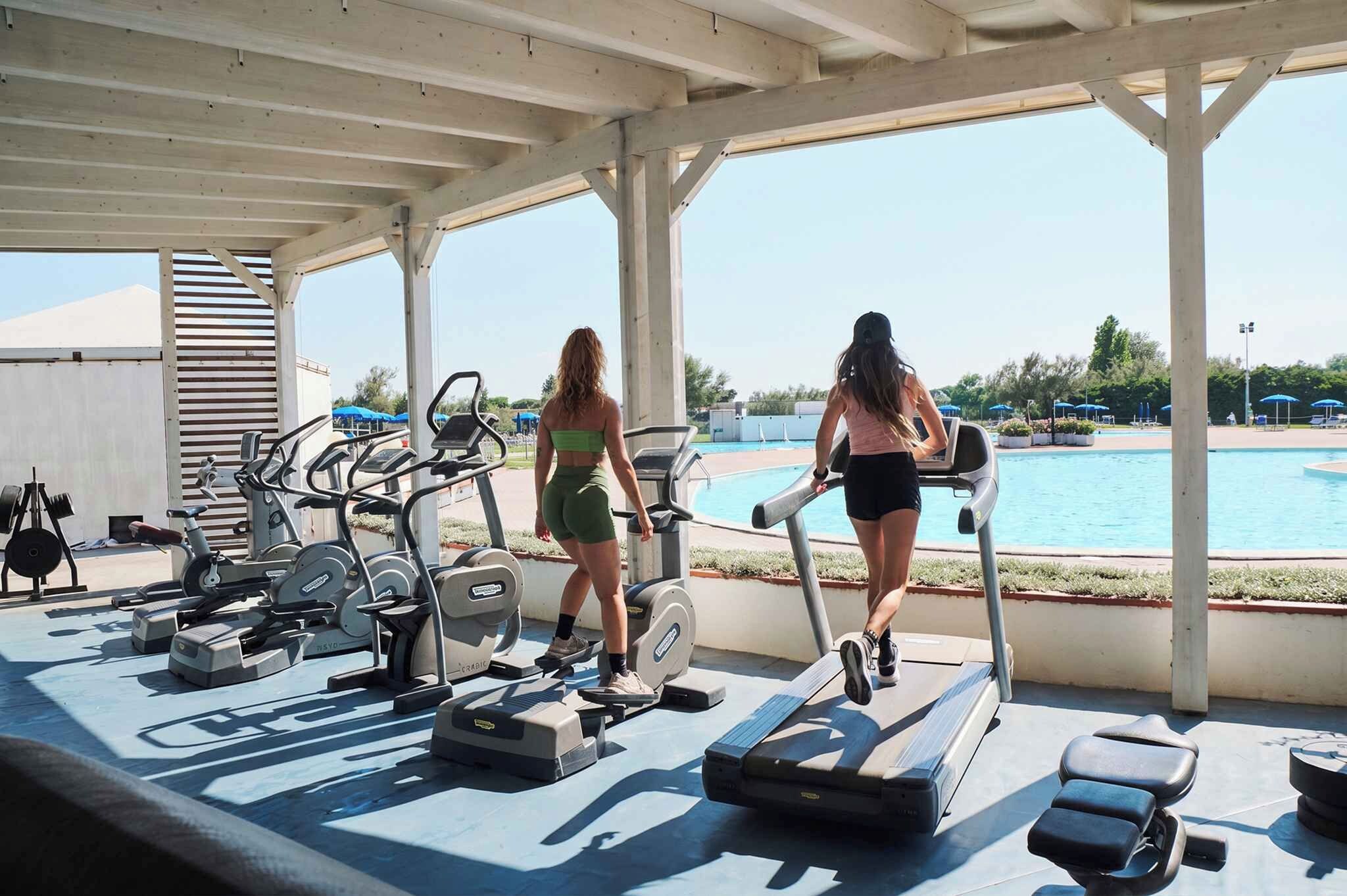 A covered outdoor gym area with exercise equipment and a pool in the background.