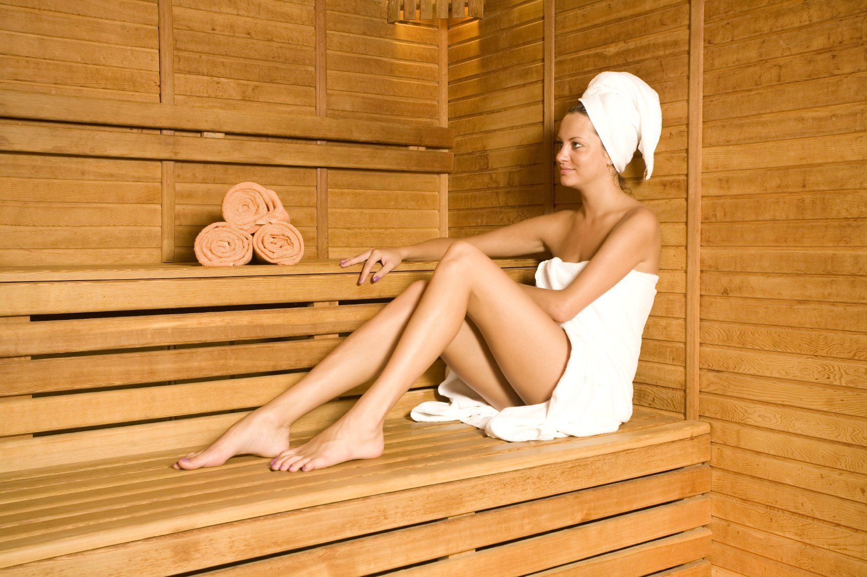 A person sitting in a wooden sauna with towels and rolled towels on a shelf.