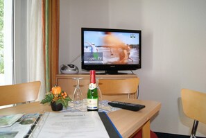 Une chambre d’hôtel avec une table en bois, un téléviseur diffusant une scène de plage, une bouteille de champagne et une télécommande.