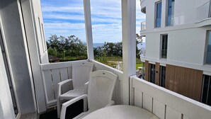 Un balcón con una silla blanca y una mesita pequeña, con vistas a un edificio de múltiples ventanas y al mar.