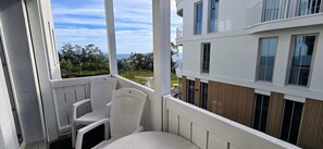 A balcony with a white chair and a small table, overlooking a building with multiple windows and a view of the sea.