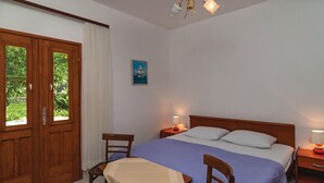 Une chambre à coucher avec un lit, deux tables de chevet, une chaise, une table recouverte d’une nappe, un tableau au mur et une porte donnant sur un paysage verdoyant.