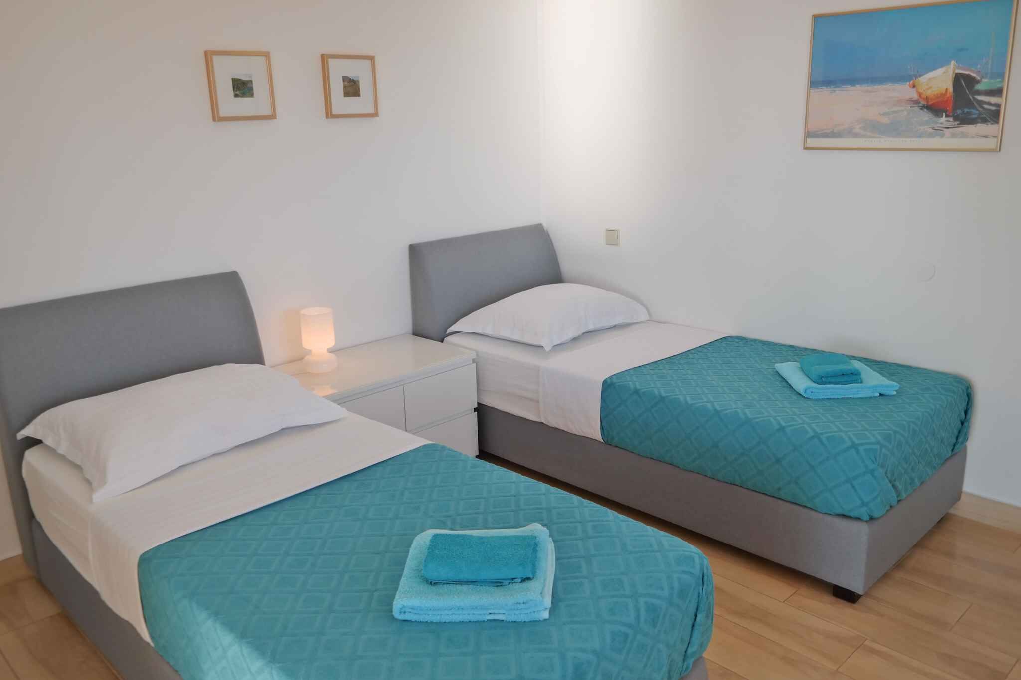 A room with two single beds, each with a grey headboard and white bedding. There is a small bedside table with a lamp on one side. The room has a light-colored wall with two framed pictures and a painting depicting a beach scene.