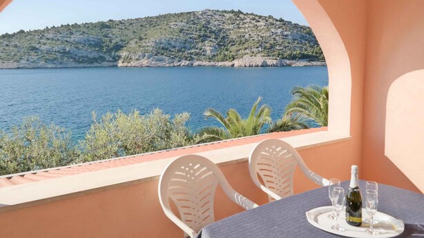A balcony with a table set for two, overlooking a body of water and a hilly landscape.