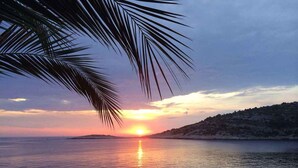 A sunset over a calm sea with boats, a rocky coastline, and a palm tree.