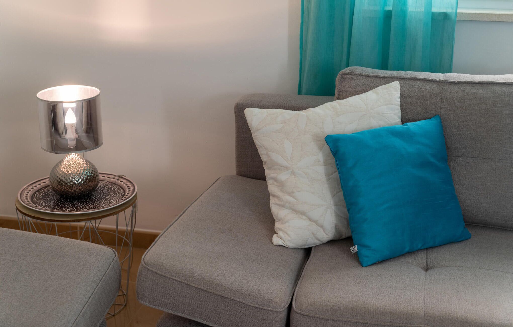 A grey sofa with a white and blue pillow, a small table with a lamp, and teal curtains.