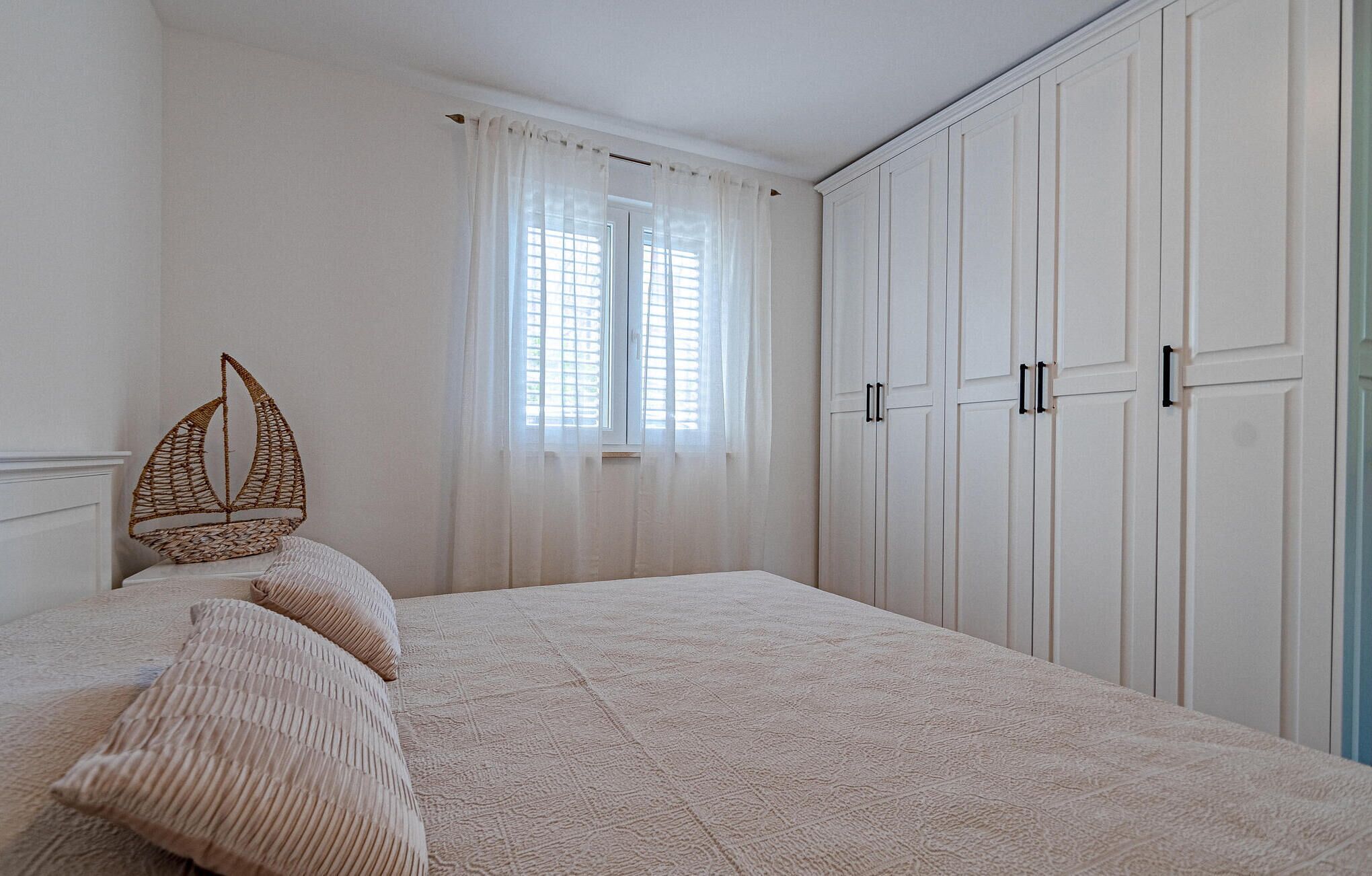 Une chambre à coucher avec un grand lit, des murs blancs, une fenêtre avec des rideaux légers et une armoire aux poignées noires.