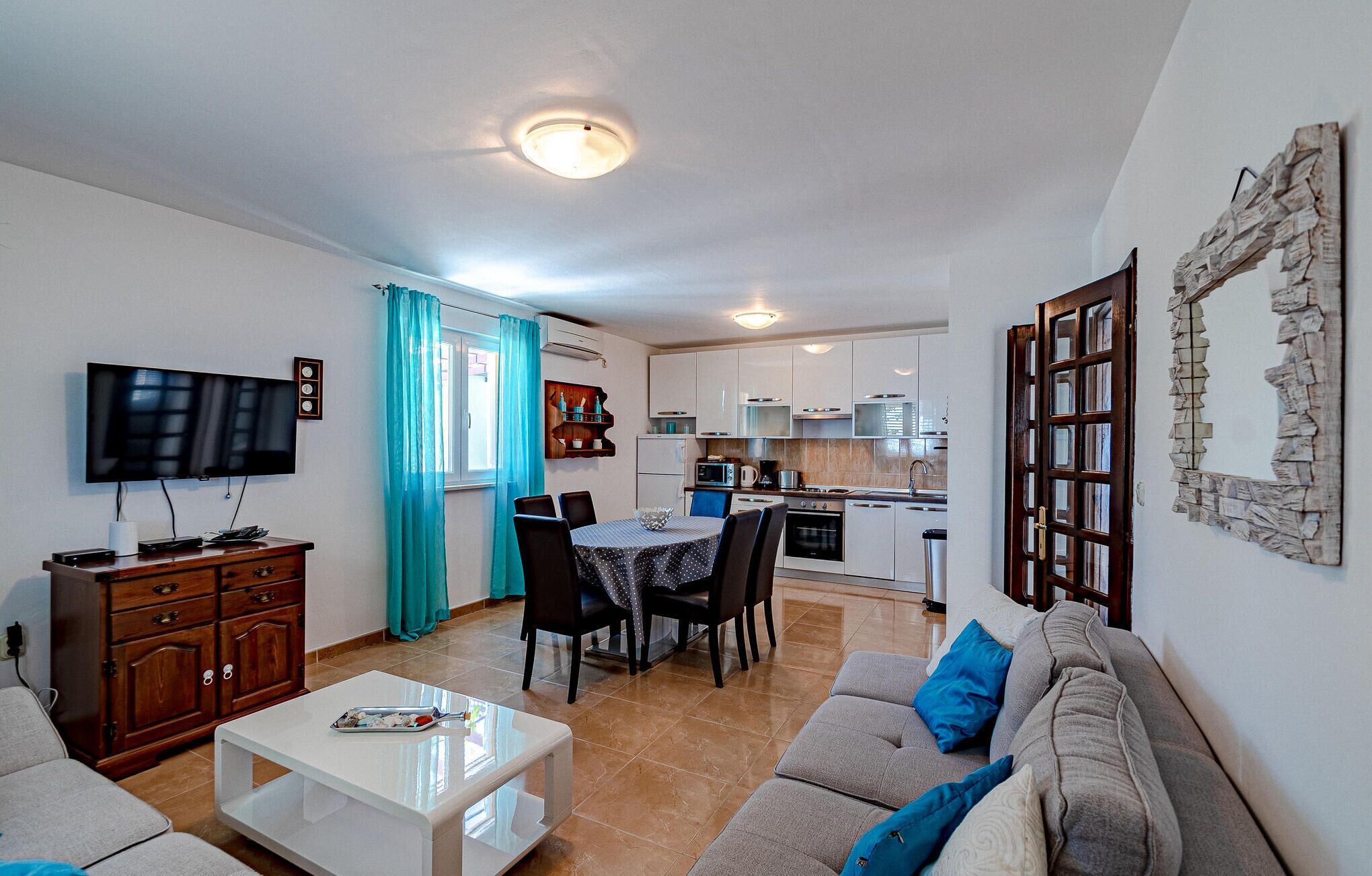 A modern living room with a flat-screen TV, a dining area with a table and chairs, a kitchen with white cabinets, and a large mirror on the wall.