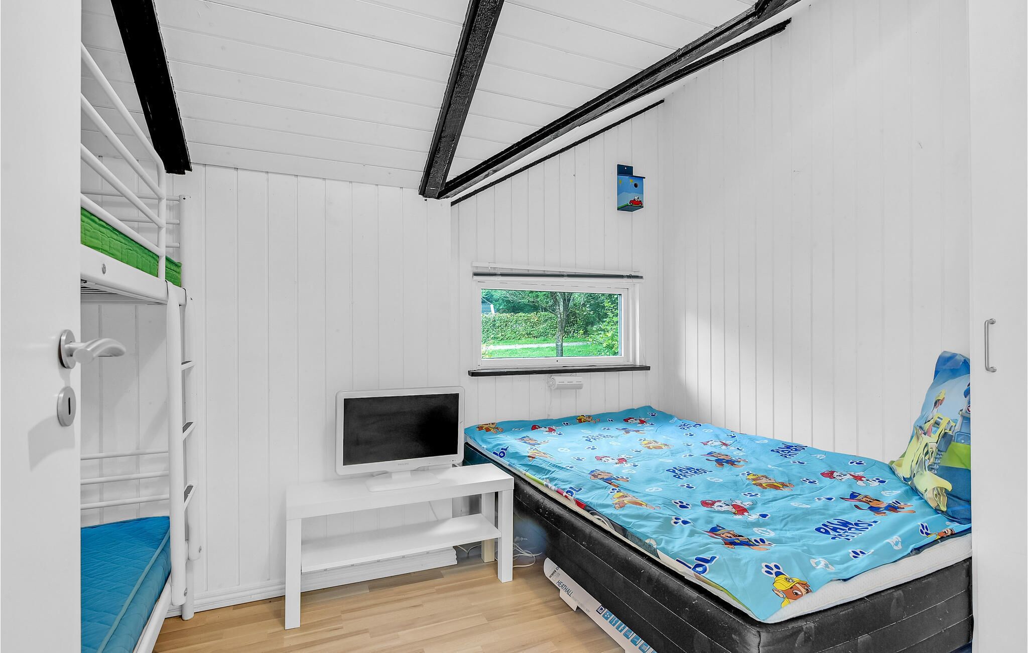 A small, well-lit room with a bed, a desk with a computer, and a window providing a view of greenery outside.