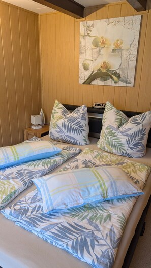 A hotel room with a bed, tropical leaf-patterned pillows, a nightstand with a lamp, and a wall-mounted painting of flowers. - Family Family Gießmann - Family Family Gießmann (Zempin)