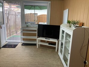 A room with a TV on a white stand, a cabinet with glass doors, a welcome mat, and a view through a door with a wooden fence.