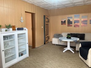 A living room with a wooden paneled wall, a white cabinet with glass doors, a beige sofa, a round table with a patterned tablecloth, and three sunset paintings on the wall.