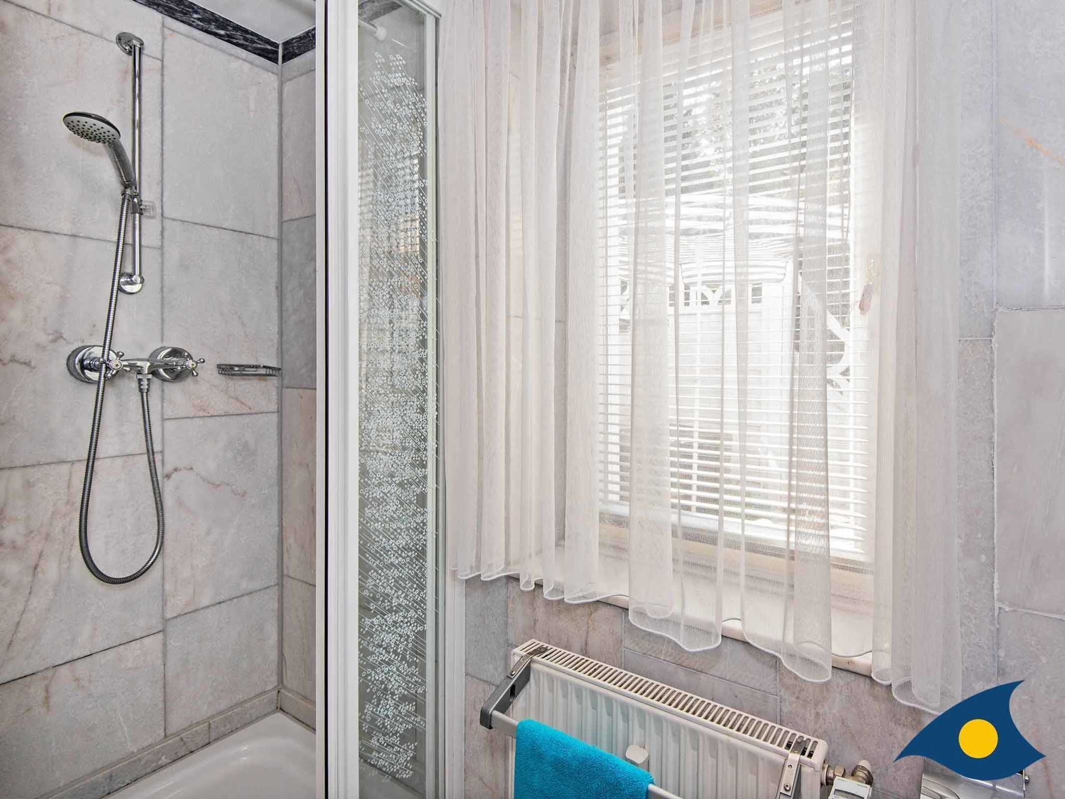A bathroom with a shower, a glass door, a window with sheer curtains, and a towel rack with a blue towel.