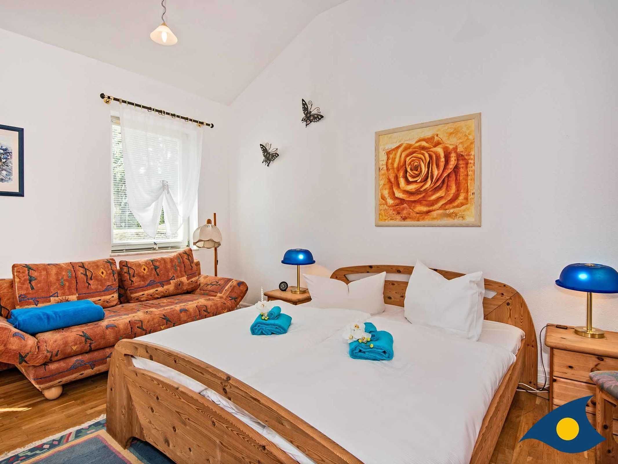 A bedroom with a wooden bed, a sofa, a window with curtains, a wall with a rose painting, and a lamp on the nightstand.