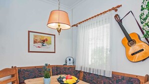 A dining area with a wooden table, chairs, a bowl of fruit, a lamp, a guitar, and a window with blinds.