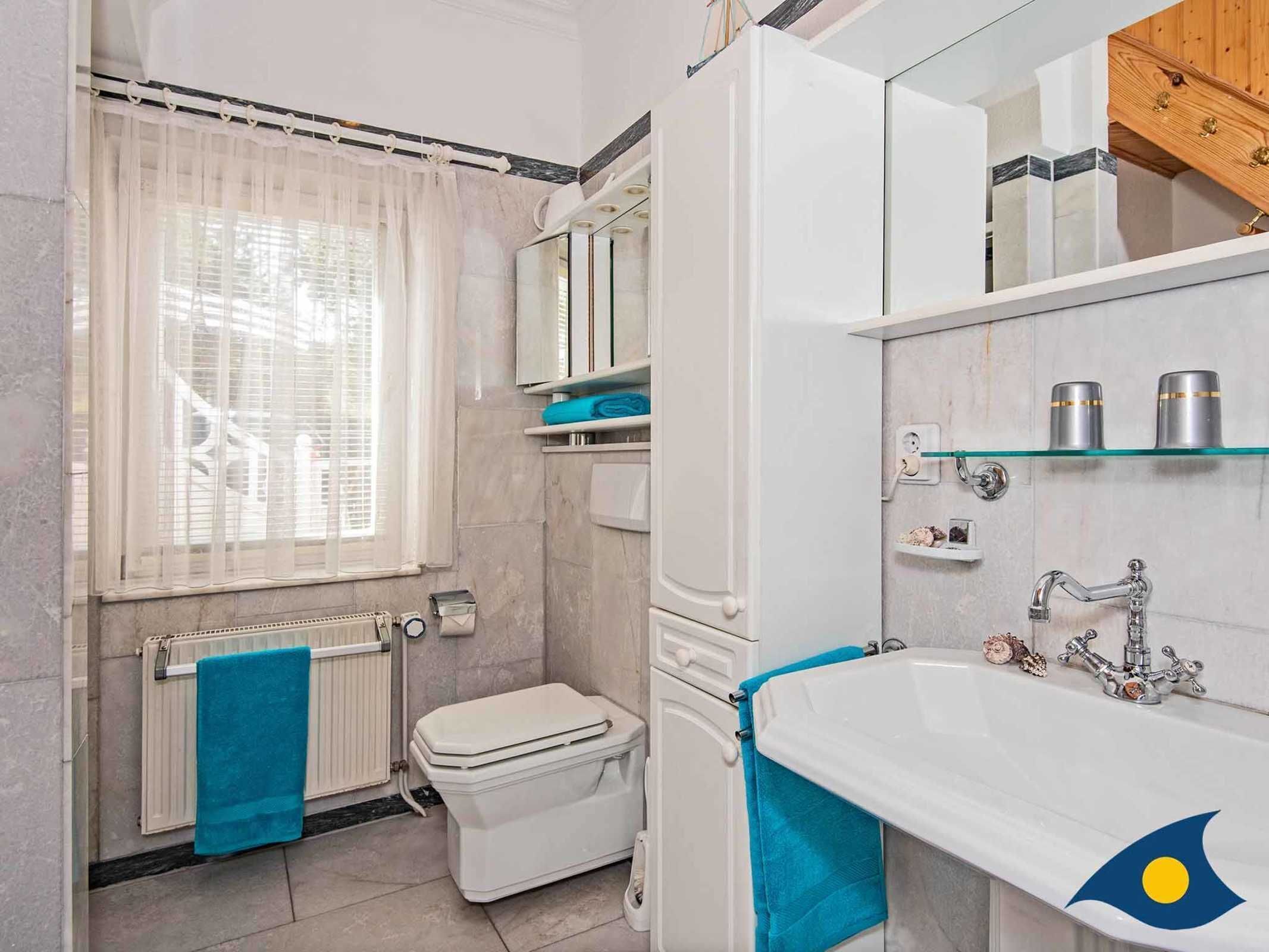 Baño con inodoro, lavabo y ducha. Hay una ventana con cortina, un perchero y un espejo sobre el lavabo.