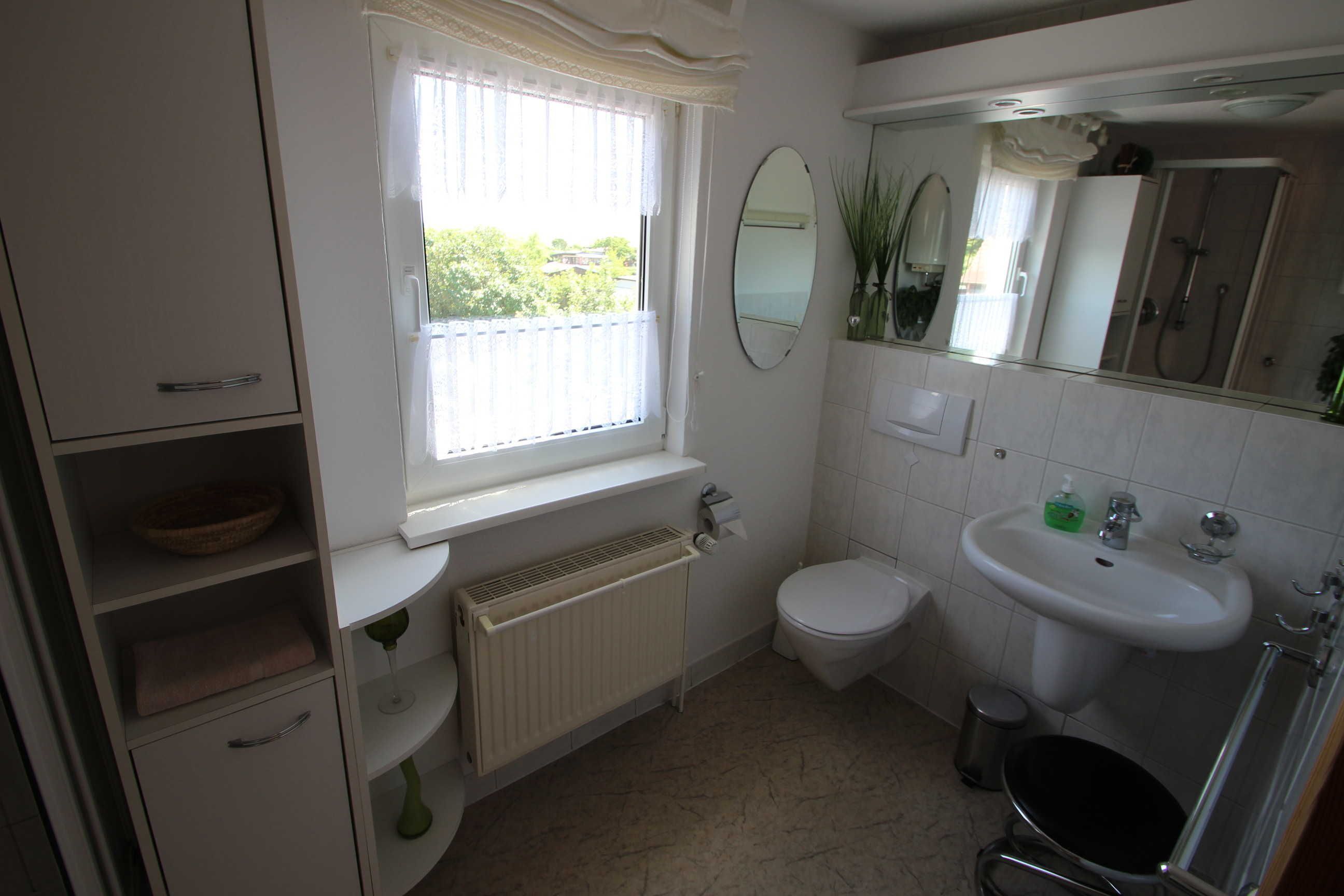 Ein Badezimmer mit Toilette, Waschbecken und Dusche. Ein Fenster mit Vorhängen, ein Heizkörper und ein Spiegel über dem Waschbecken sind vorhanden.