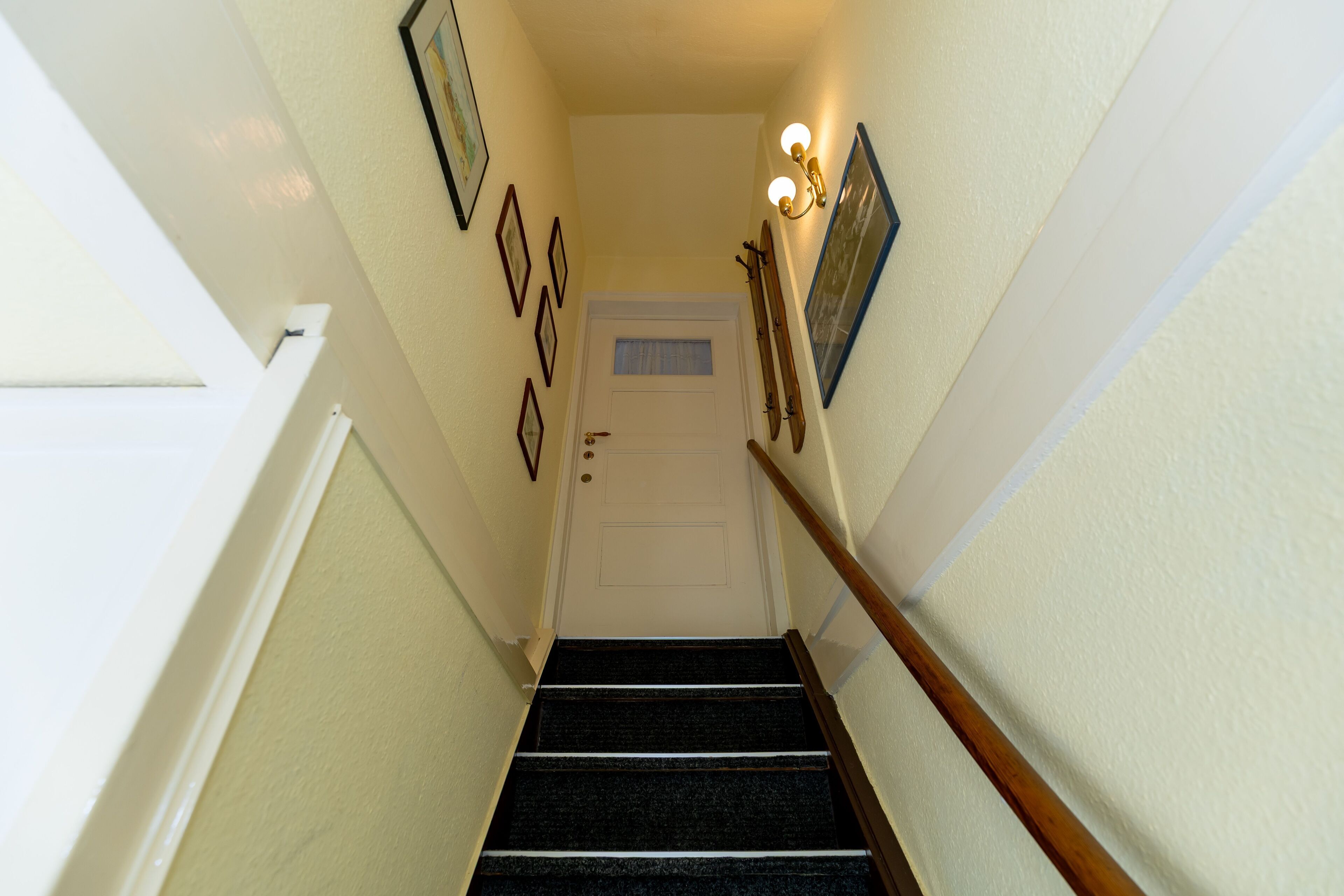 Un escalier surmonté d’une porte blanche, avec une main courante sur le côté droit et des tableaux encadrés accrochés aux murs.