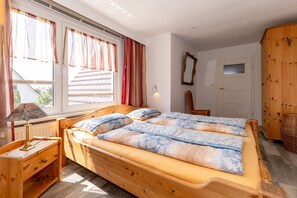 Een houten bed met een blauw-beige dekbedovertrek, een nachtkastje met een lamp en een raam met gordijnen.