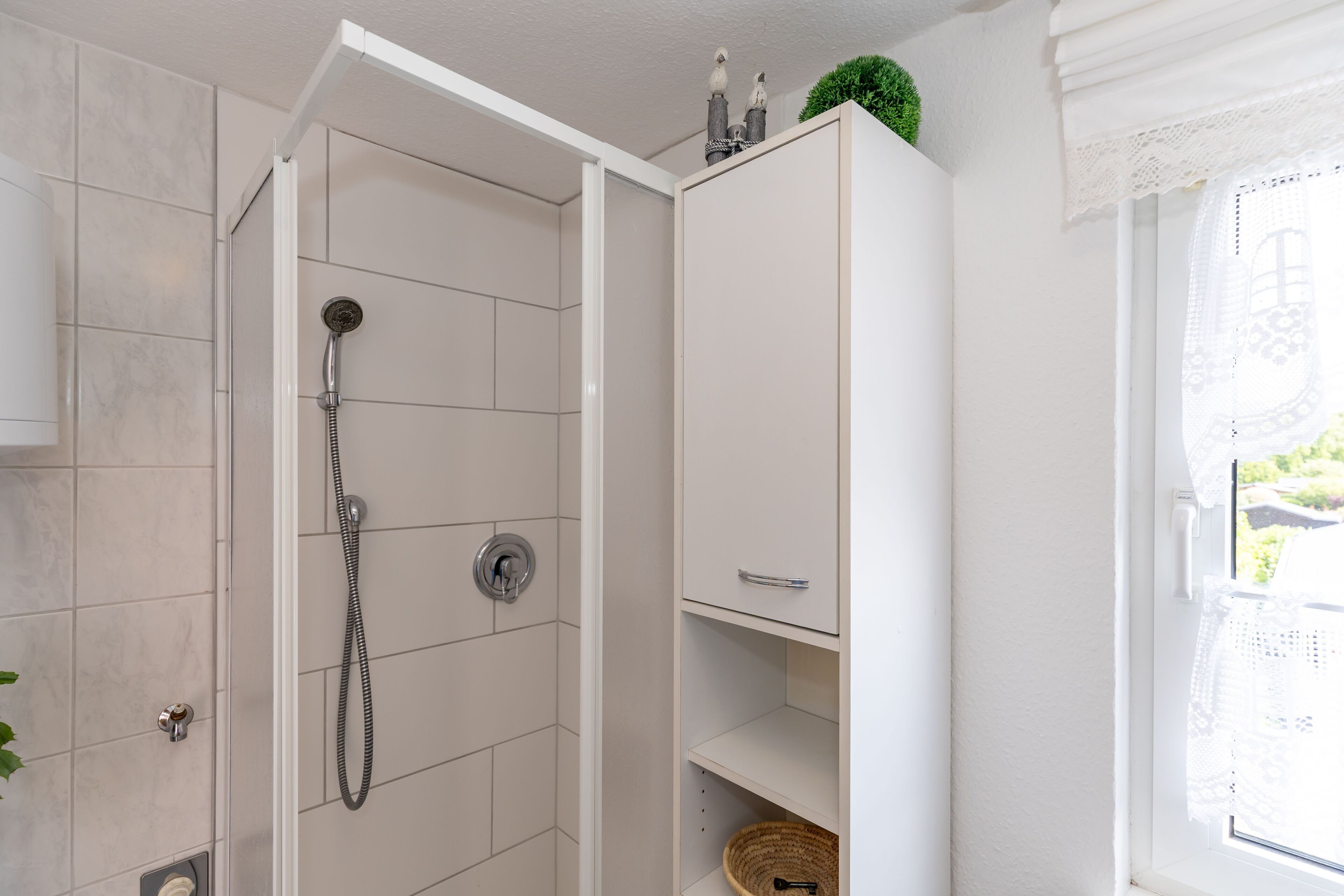 Une salle de bain avec une douche, un meuble blanc doté d’une poignée et une fenêtre ornée de rideaux en dentelle.