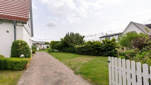 A paved pathway leading to a white house with a red roof, surrounded by greenery and a white picket fence. - Apartment garden view - 2-room apartment garden view (Rerik)