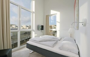 A modern hotel room with a large bed, a TV, a desk, and a view of the city through large windows. - Dünenvilla 10, Whg 5 (Travemünde-Priwall)
