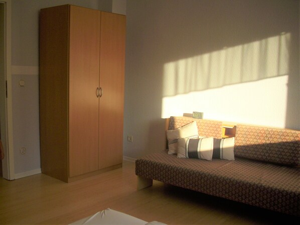 A room with a wooden wardrobe, a patterned sofa with cushions, and a window casting shadows on the wall.