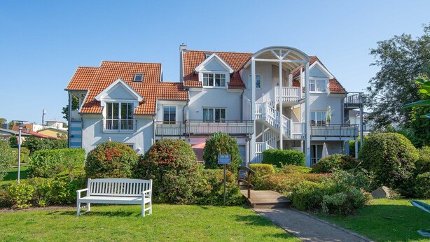 A multi-story residential building with a red-tiled roof, balconies, and a garden area with a bench.
