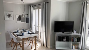 A modern living room with a dining area, a flat-screen TV on a stand, and a bookshelf.