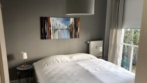 A modern bedroom with a large bed, a small round table, a lamp, a radiator, and a view of greenery outside.