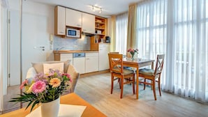 A modern kitchen with built-in appliances, a dining area with wooden furniture, and large windows with sheer curtains. - Villa Aquamarina Whg. 1, SOUTH FACING BALCONY, CHAIR - Villa Aquamarina, 1st ROW, partly. (Heringsdorf)