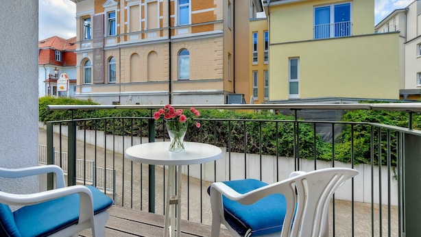 A balcony with a white table and chairs, a vase of flowers, and a view of multi-story buildings. - Villa Aquamarina Whg. 1, SOUTH FACING BALCONY, CHAIR - Villa Aquamarina, 1st ROW, partly. (Heringsdorf)