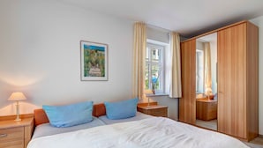 A bedroom with a large bed, two bedside tables with lamps, a wardrobe, a window with curtains, and a painting on the wall. - Villa Aquamarina Whg. 1, SOUTH FACING BALCONY, CHAIR - Villa Aquamarina, 1st ROW, partly. (Heringsdorf)