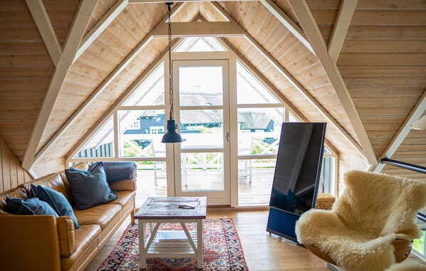 A cozy loft bedroom with a sloped ceiling, a large window, a sofa, a coffee table, a chair, and a TV.
