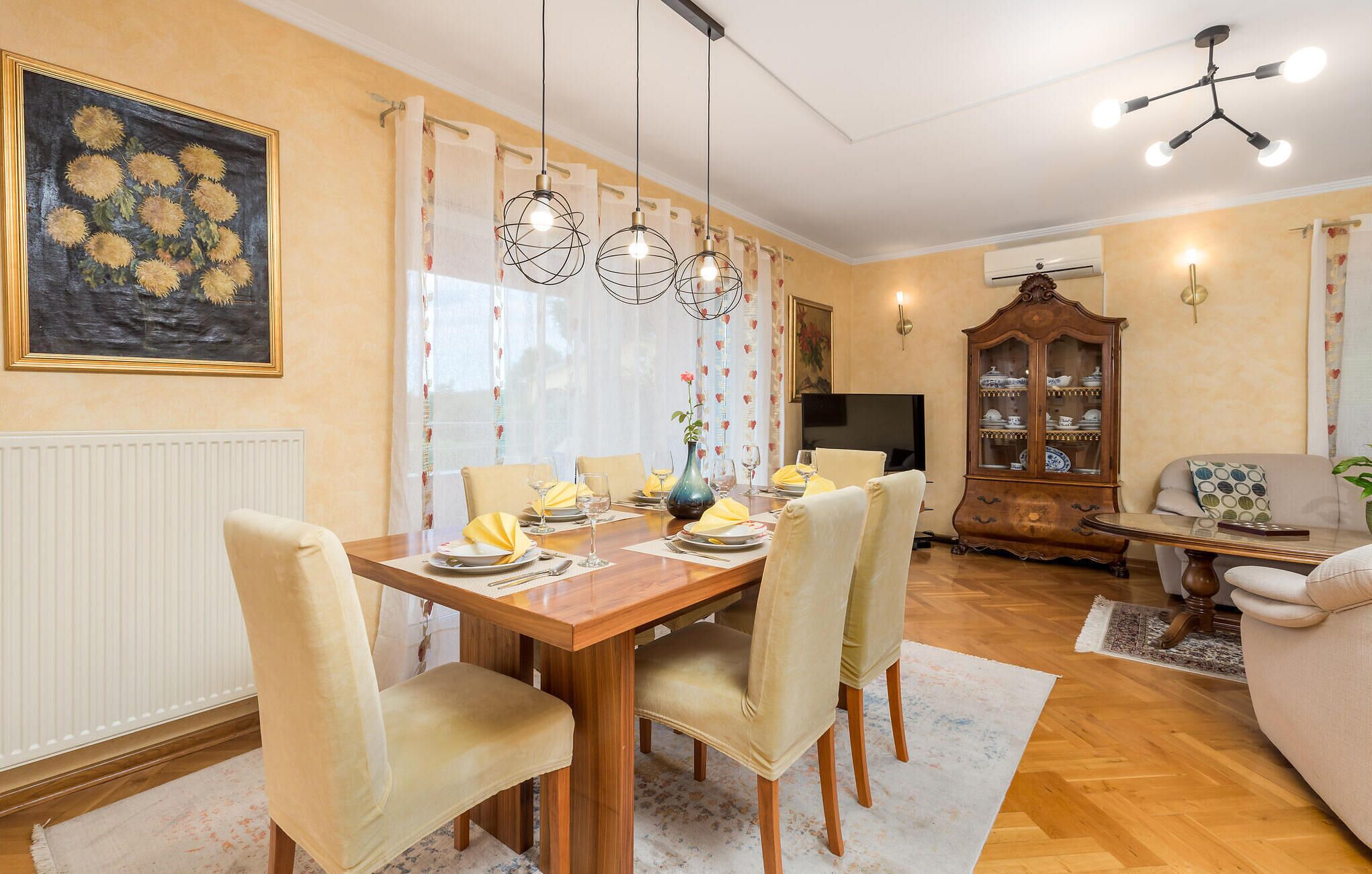 A dining area with a wooden table set for four, surrounded by chairs. There is a cabinet with dishes and a television in the background.