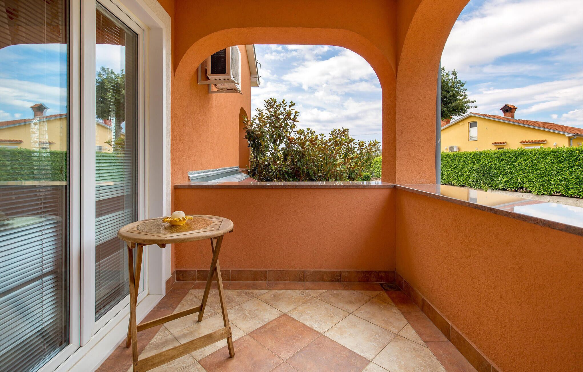 Ein Balkon mit einem kleinen Tisch, ein Fenster mit Jalousien und Blick auf ein gelbes Gebäude und Grünflächen.