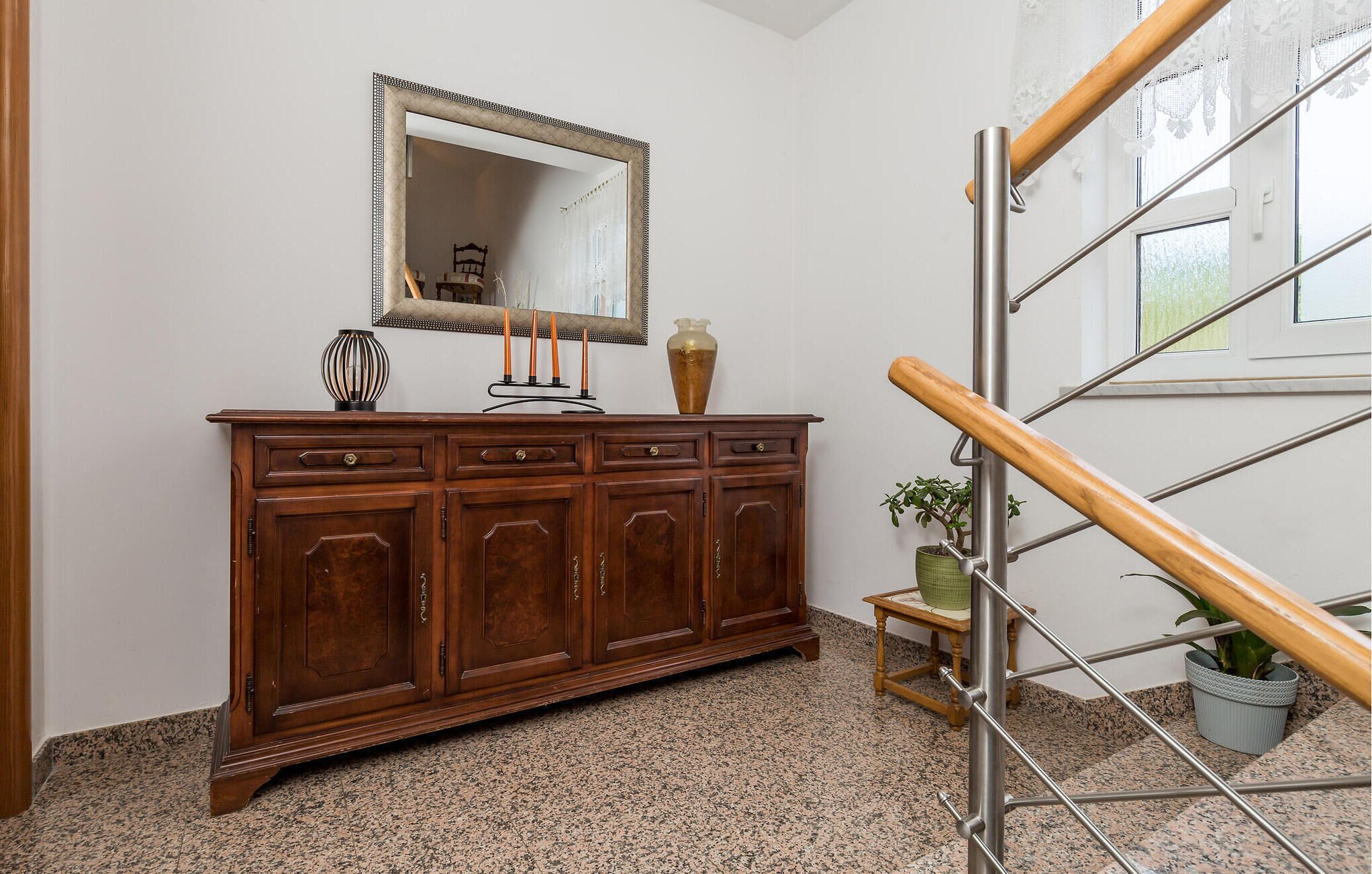 A wooden cabinet with a mirror above it, a vase, and a candle holder. There is a staircase with a wooden handrail and metal balusters.