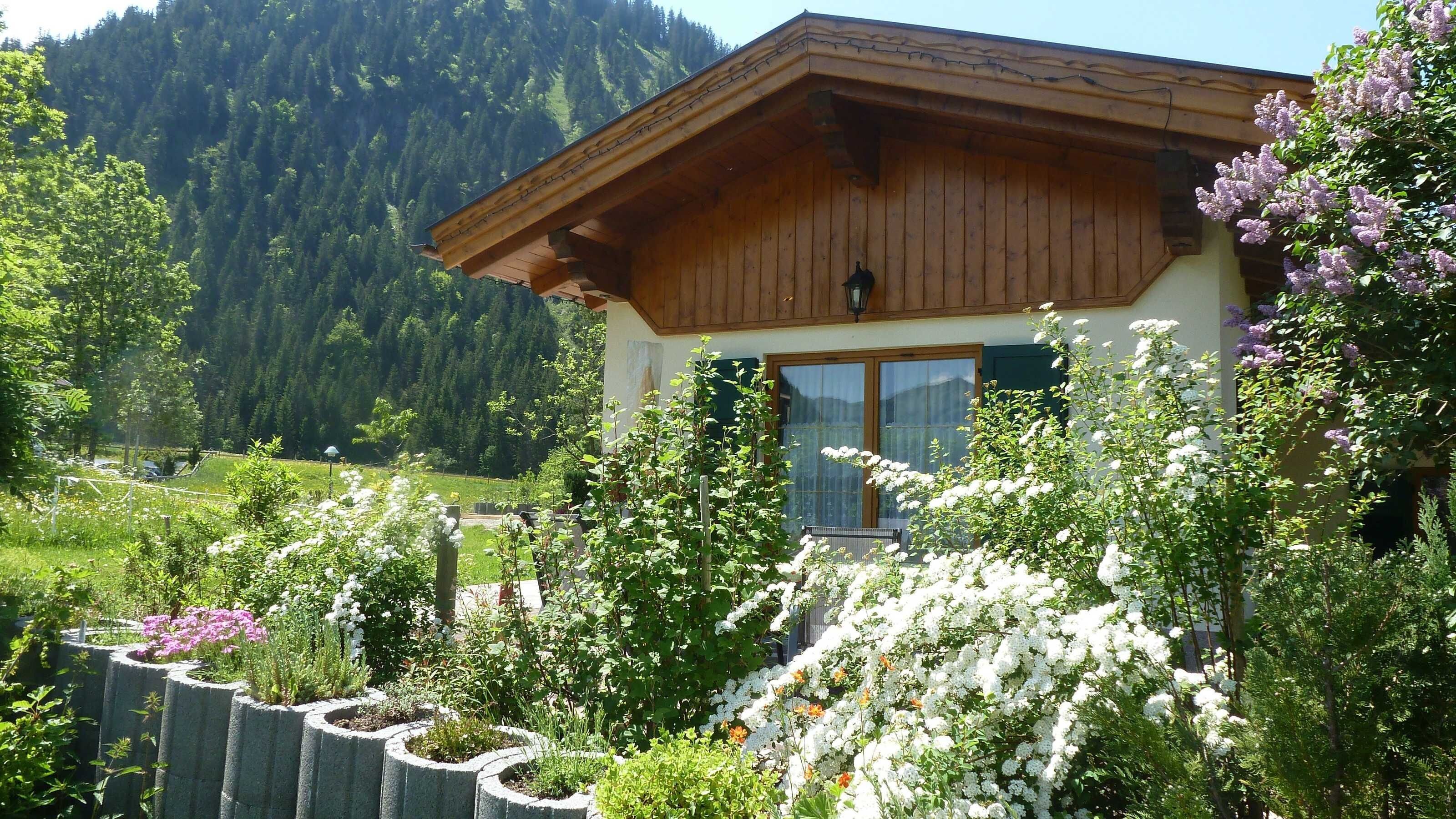 A wooden chalet with a sloped roof, surrounded by lush greenery and flowering plants.