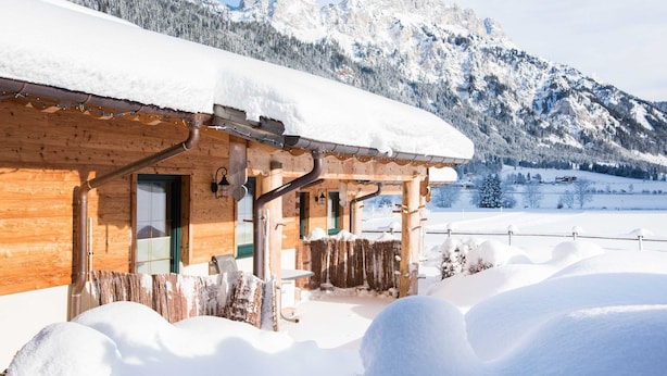Eine Holzhütte mit verschneitem Dach, einer hölzernen Veranda und Blick auf verschneite Berge im Hintergrund.
