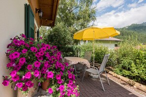 A patio with a table and chairs, a yellow umbrella, and a potted plant with pink flowers. - Chalet vacation home 11 - Almdorf Tirol (Haldensee)