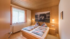 Ein Schlafzimmer mit Holzwänden, einem Bett, einem Fenster mit Vorhängen, einem Heizkörper und einem an der Wand hängenden Bild.