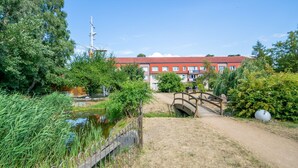 A building with a red roof and multiple windows, surrounded by greenery and a small bridge over a pond.