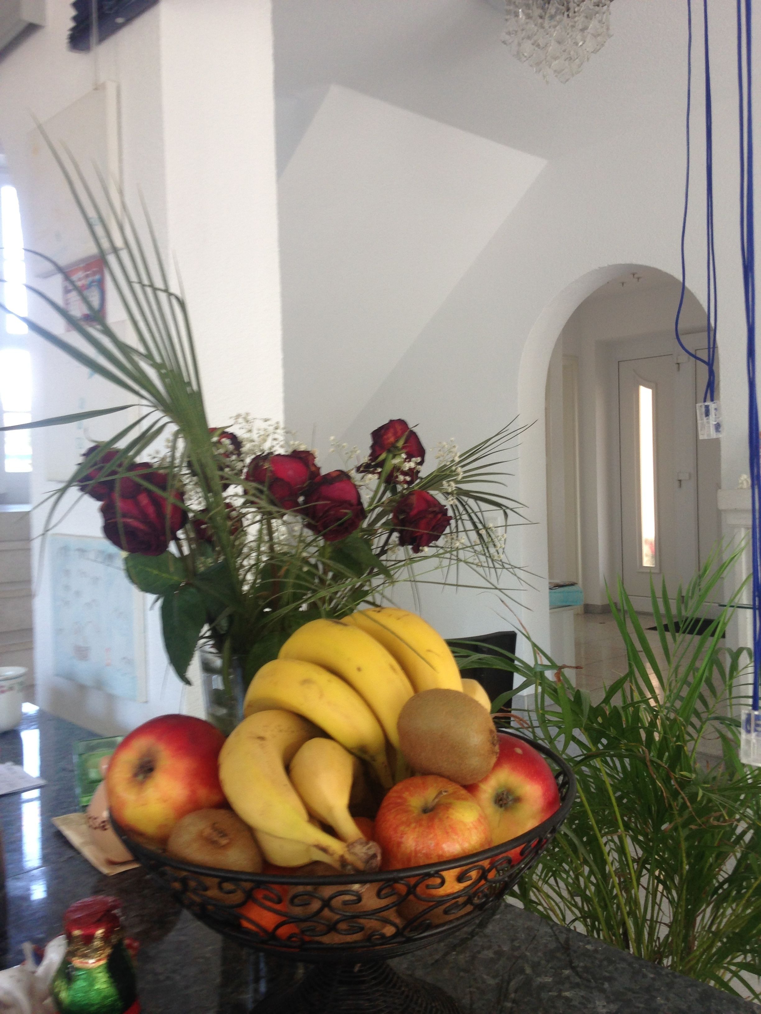 A kitchen counter with a fruit bowl containing bananas, apples, pears, and kiwis. There is a vase with red roses and other flowers.