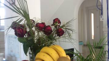 A kitchen counter with a fruit bowl containing bananas, apples, pears, and kiwis. There is a vase with red roses and other flowers.
