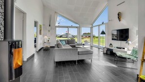 A modern living room with a fireplace, large windows, a flat-screen TV, and a grey sectional sofa. - Cozy home in Sæby with house sea view (Sæby)