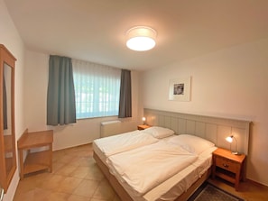 A bedroom with two beds, a window with curtains, a bedside table with a lamp, and a radiator.